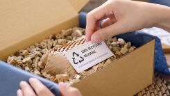 A shipping box filled with reusable packing material, with a hand visible holing a small card that reads "Reusable, Recyclable"