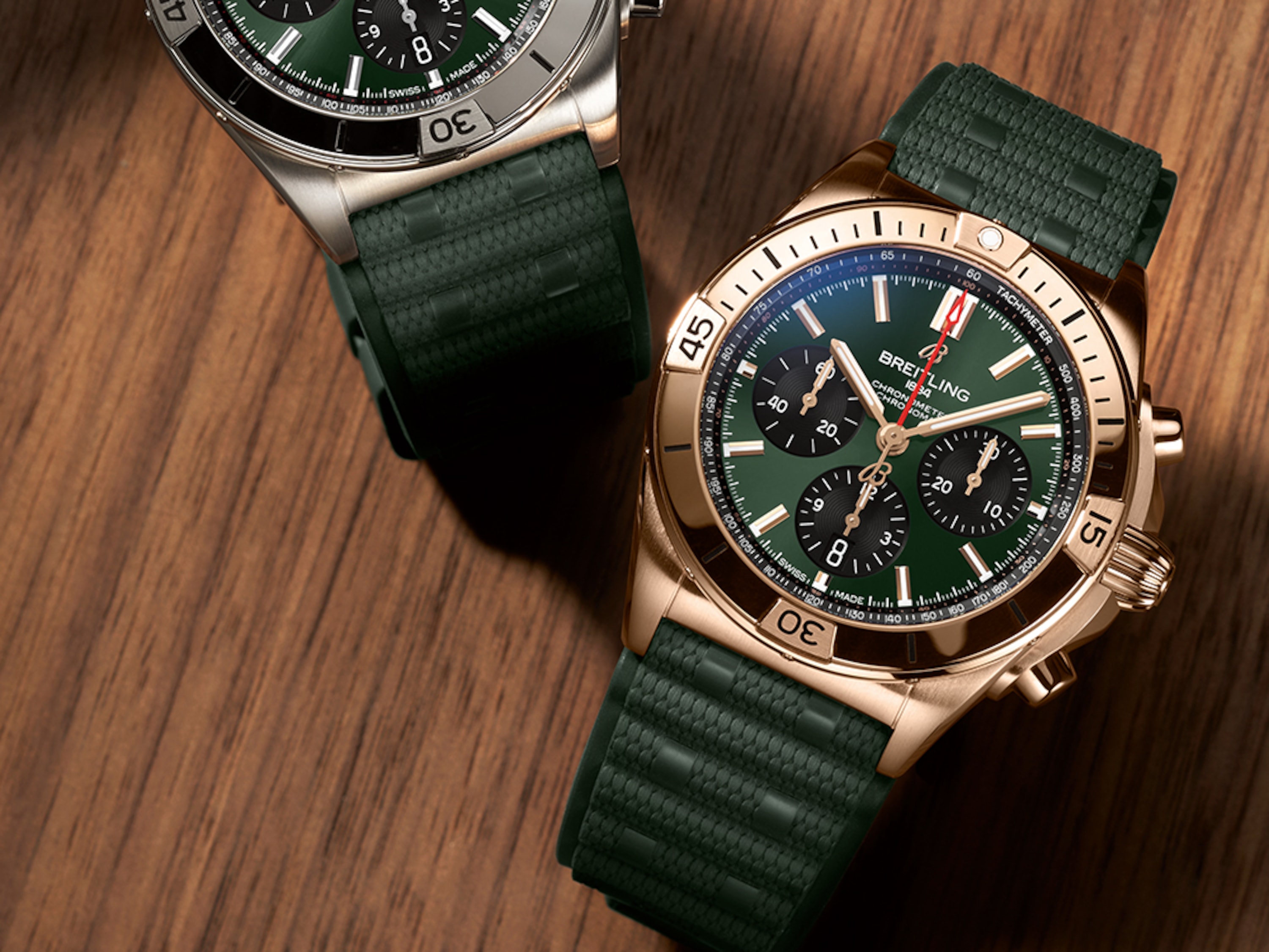 A gold and a silver Chronomat with dark green rubber straps on a brown table.