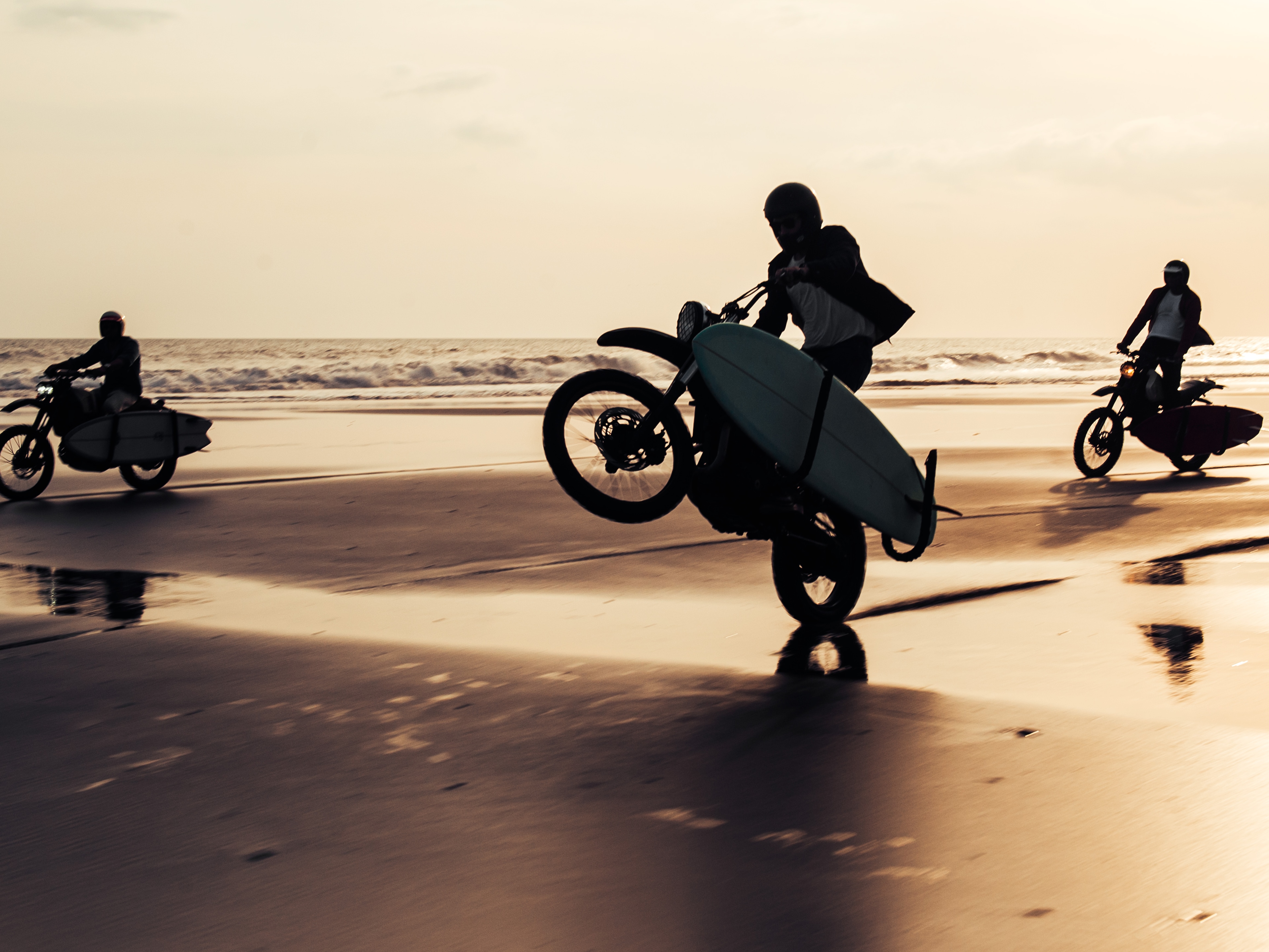 Three Deus Ex Machina motorcycles with surfboards in tow ride through the surf on the beach at sunset 