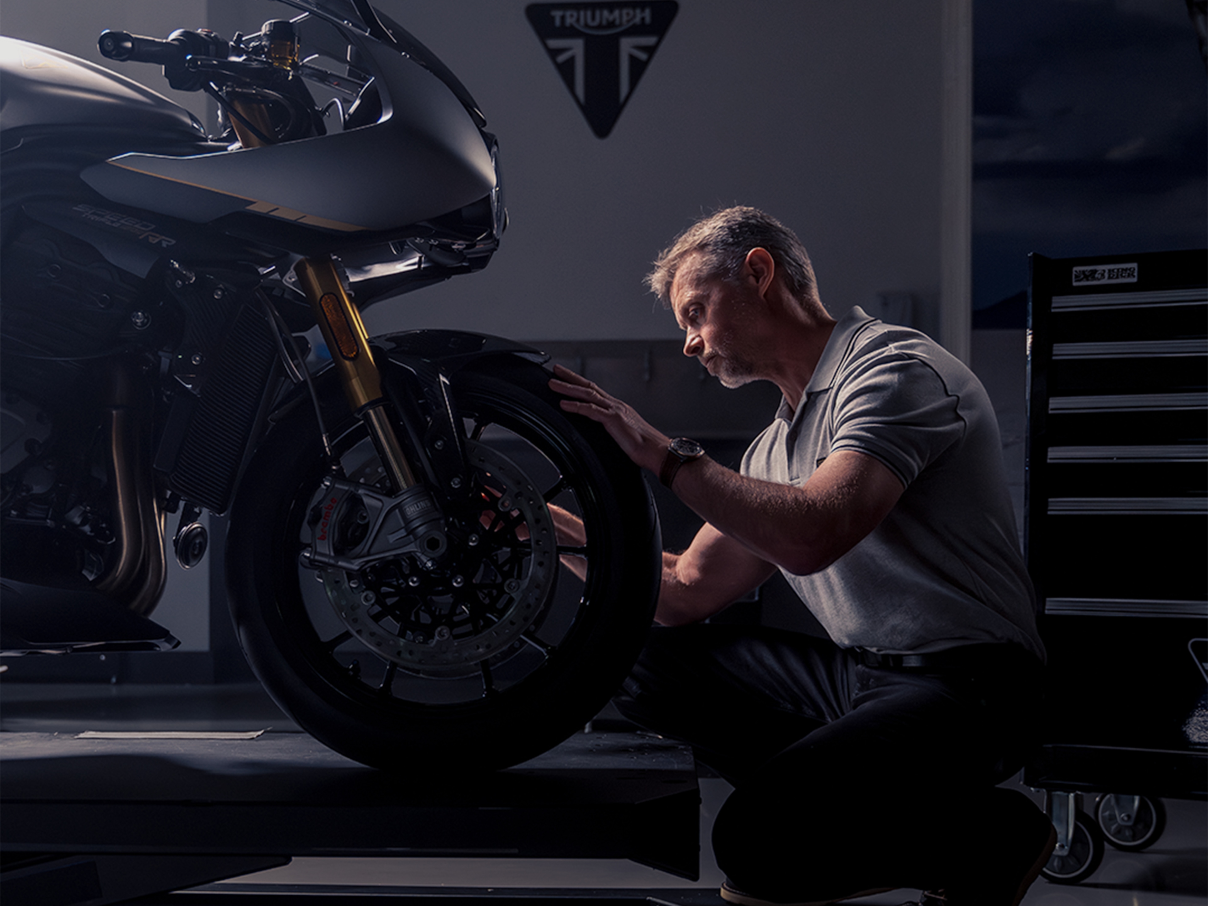 Man with Breitling watch in focus kneels in front of Triumph motorcycle